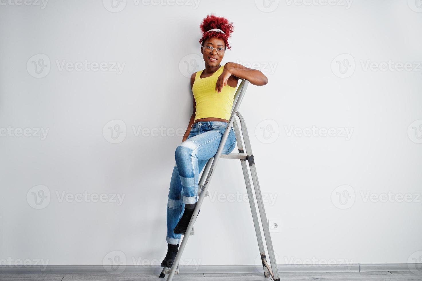 African american woman in yellow singlet and eyglasses against white wall stand om iron ladder. photo