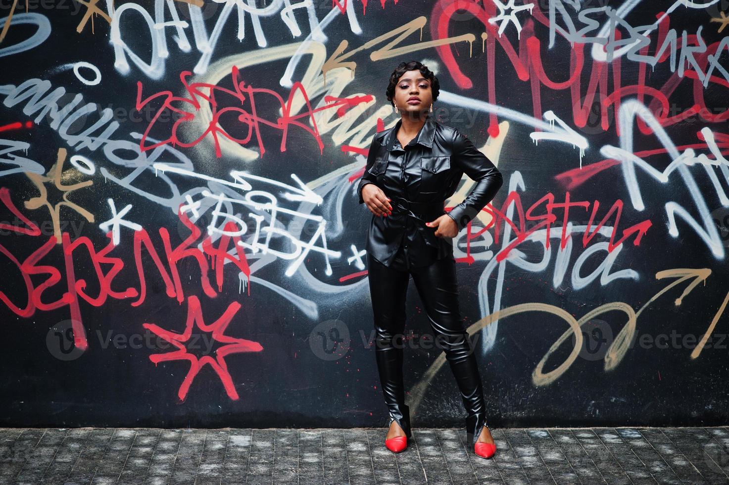 hermosa mujer afroamericana de moda posando en chaqueta de cuero negro y pantalones en la calle contra la pared de graffiti. foto