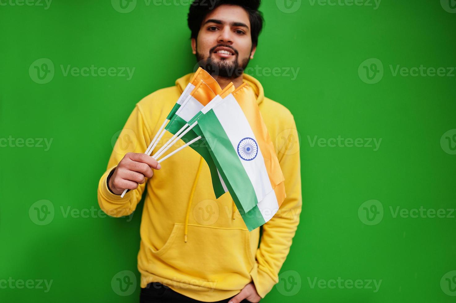Urban young hipster indian man in a fashionable yellow sweatshirt hold flags of India in hands. Cool south asian guy wear hoodie isolated on green background. photo