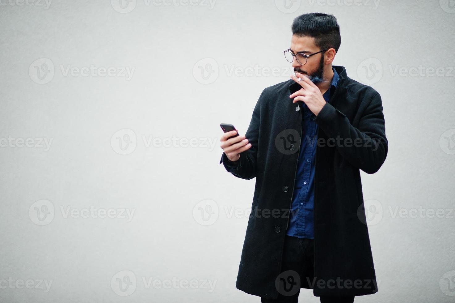 Middle eastern entrepreneur wear black coat and blue shirt, eyeglasses standing against white wall, smoking cigarette and looking on phone. photo