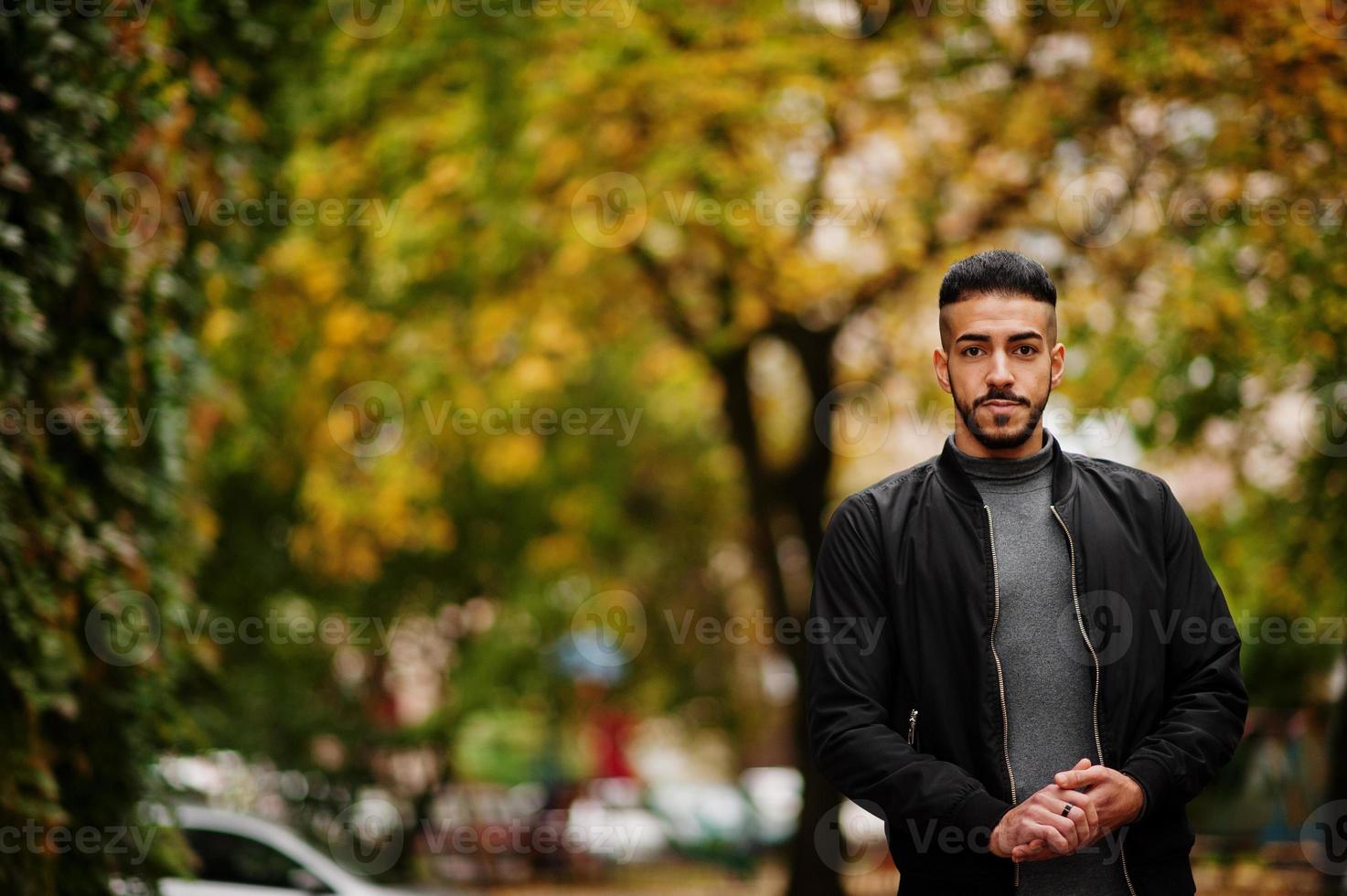 Portrait of stylish arab beard man wear grey turtleneck and black jaket. Arabian model guy on a background of autumn leaves. photo