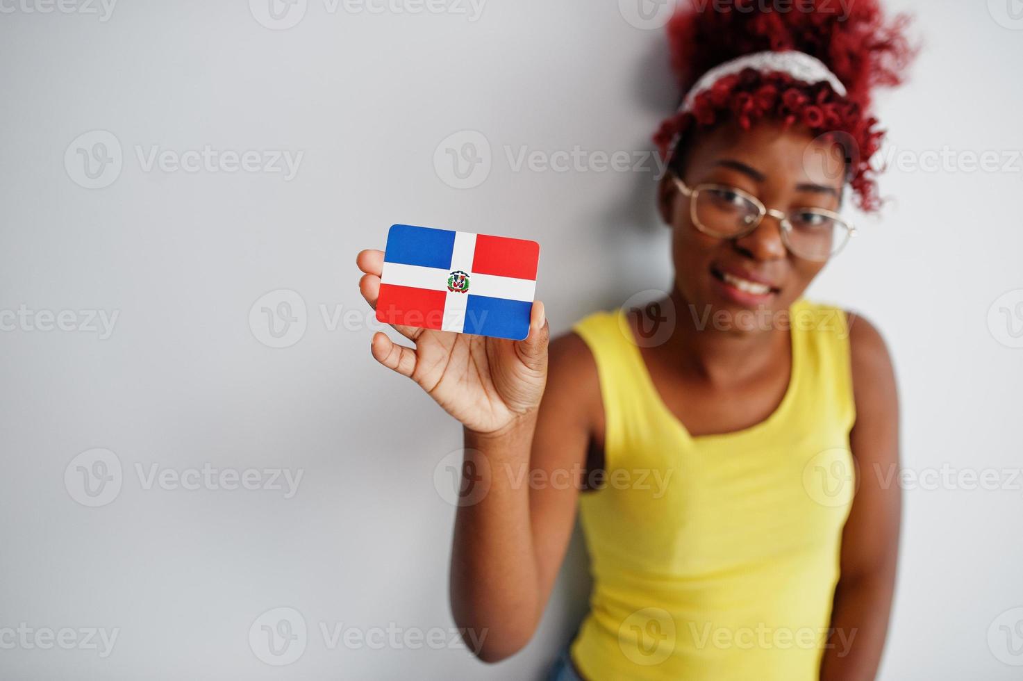 mujer afroamericana con cabello afro, use camiseta amarilla y anteojos, sostenga la bandera de la república dominicana aislada sobre fondo blanco. foto
