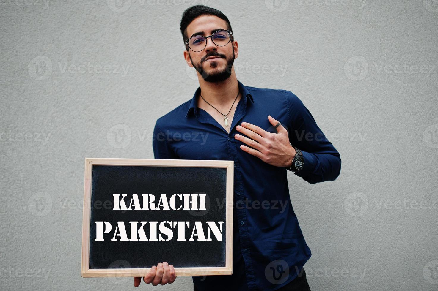 el hombre árabe usa camisa azul y anteojos sostiene una tabla con la inscripción karachi pakistán. ciudades más grandes en el concepto del mundo islámico. foto