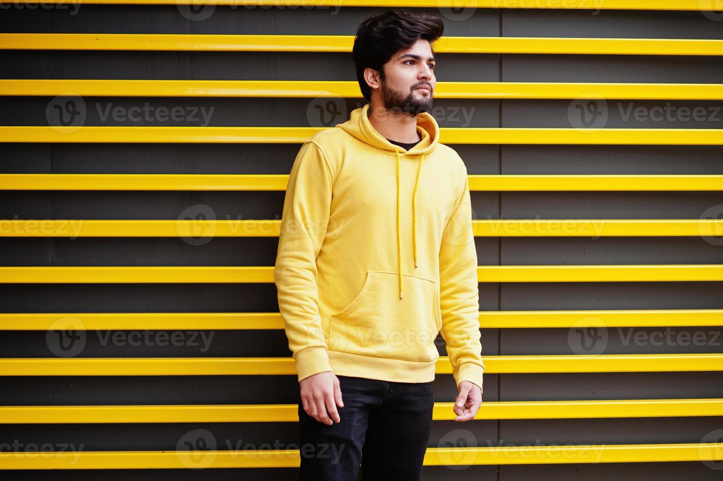 hombre indio joven hipster en una sudadera amarilla de moda. un chico fresco del sur de Asia usa una sudadera con capucha contra un fondo rayado. 11014658 Foto de stock en