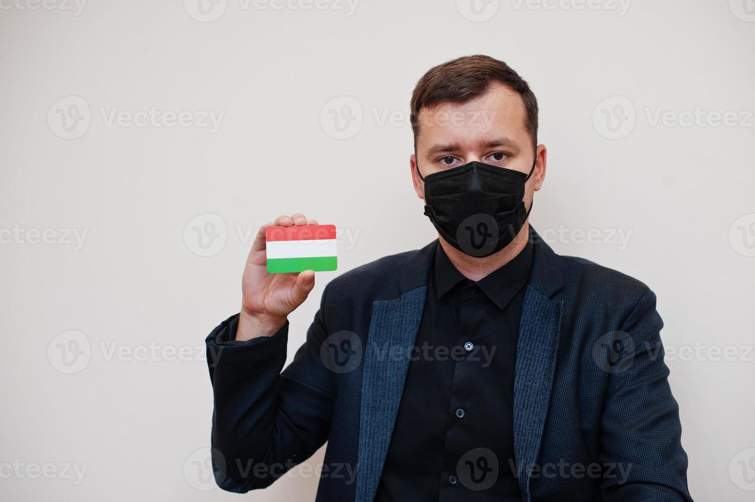 el hombre europeo usa negro formal y protege la máscara facial, sostiene la tarjeta de la bandera de hungría aislada en el fondo blanco. concepto de país covid coronavirus de europa. foto