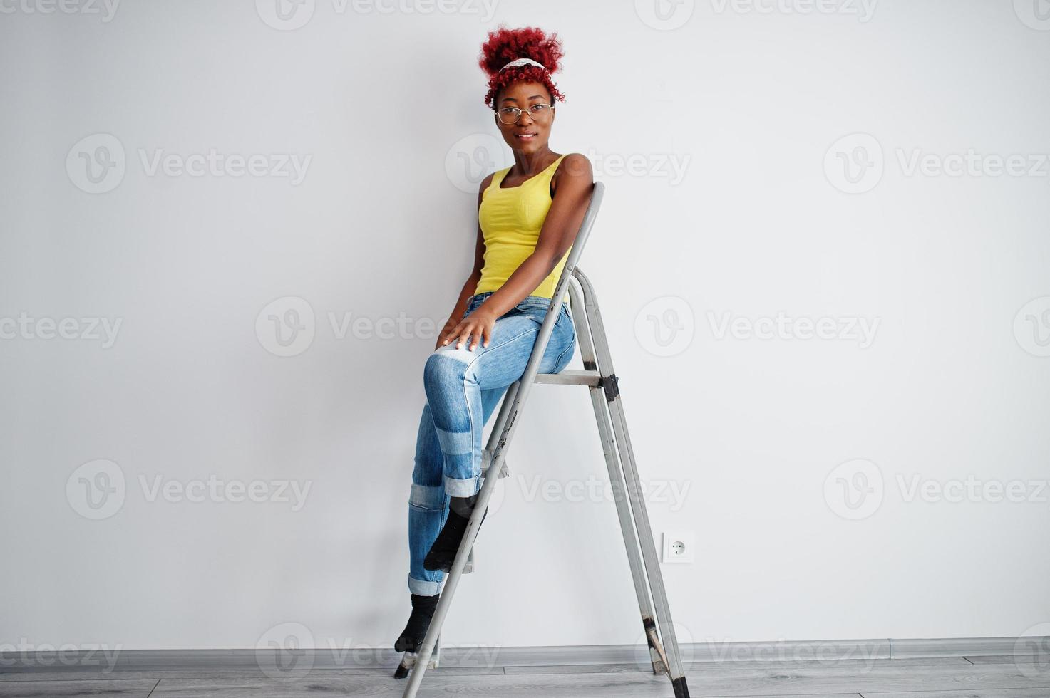 African american woman in yellow singlet and eyglasses against white wall stand om iron ladder. photo