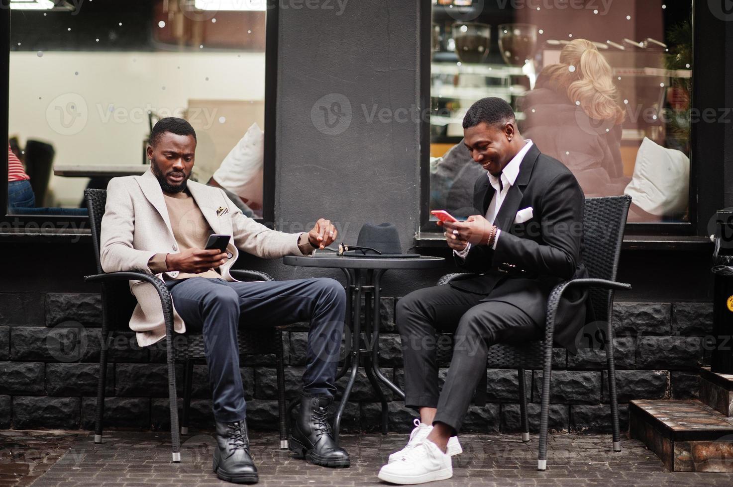 dos hombres negros de moda sentados al aire libre con teléfono móvil. retrato de moda de modelos masculinos afroamericanos. llevar traje, abrigo y sombrero. foto