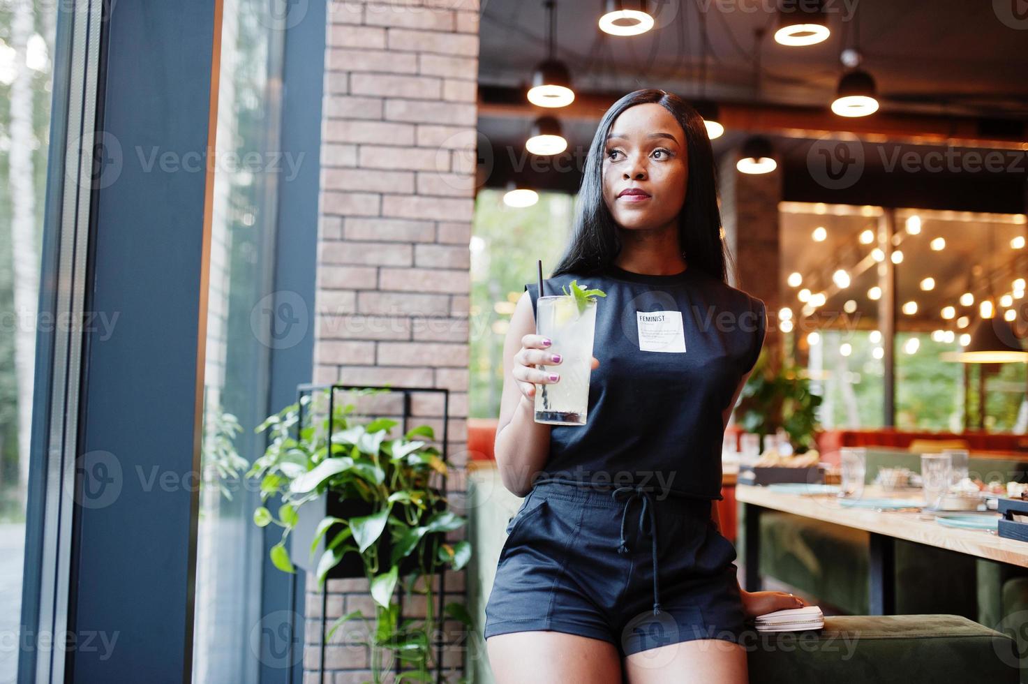 Mujer afroamericana feminista de moda vestida con camiseta negra y pantalones cortos, posada en un restaurante con vaso de limonada. foto