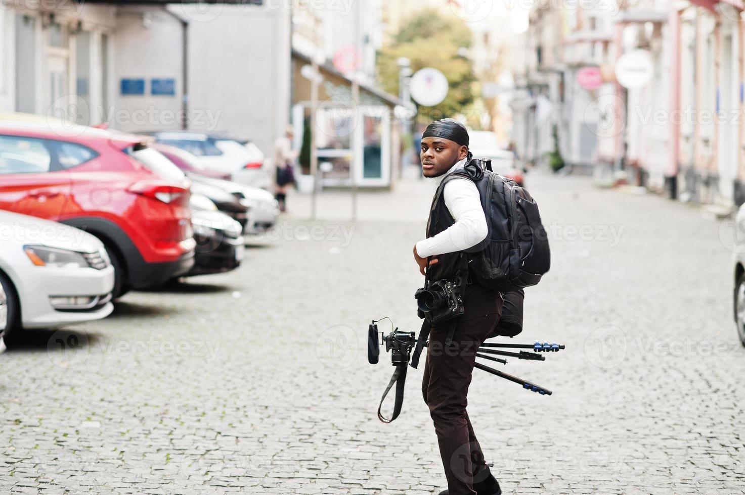 Young professional african american videographer holding professional camera with tripod pro equipment. Afro cameraman wearing black duraq making a videos. photo