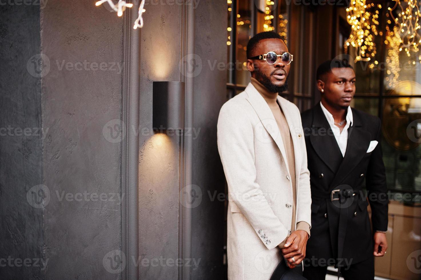 Two fashion black men pose against house with garlands. Fashionable portrait of african american male models. Wear suit, coat and hat. photo