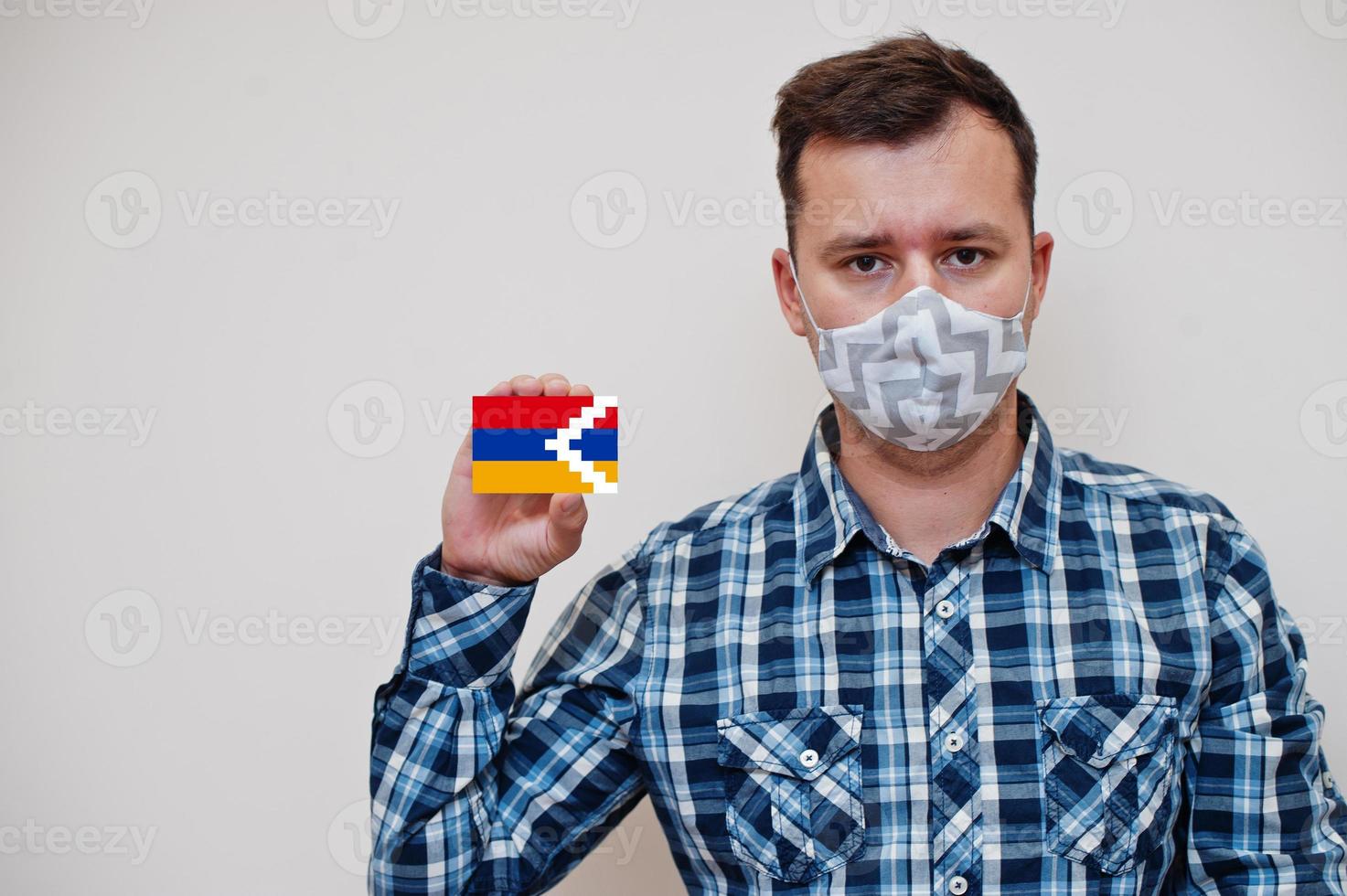 Man in checkered shirt show Artsakh flag card in hand, wear protect mask isolated on white background. Europe countries Coronavirus concept. photo
