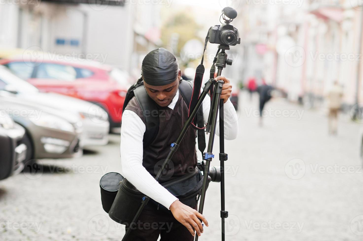 Young professional african american videographer holding professional camera with tripod pro equipment. Afro cameraman wearing black duraq making a videos. photo