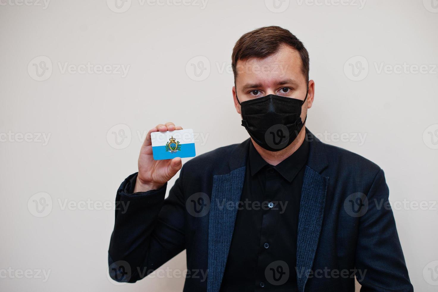 European man wear black formal and protect face mask, hold San Marino flag card isolated on white background. Europe coronavirus Covid country concept. photo