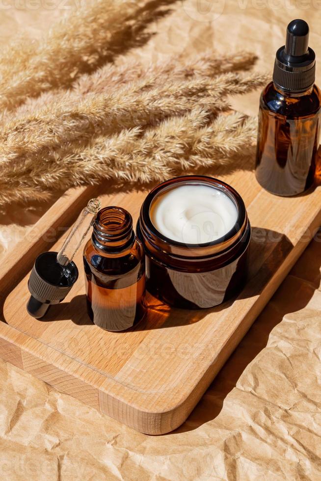 Essential oils with droppers pipette and cream on wooden tray on beige background. Dry ear reeds or pampas grass decoration. Professional bottle for facial and body treatment. Unbranded package photo