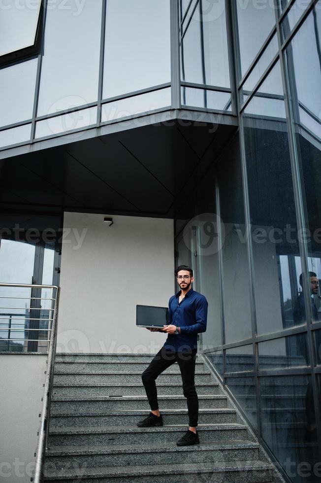 Middle eastern entrepreneur wear blue shirt, eyeglasses with laptop against office building. photo