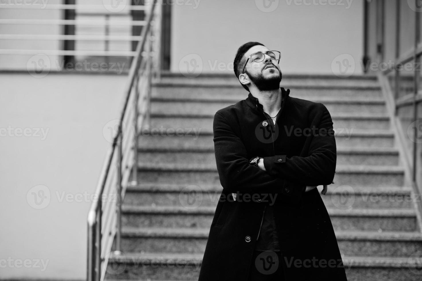 empresario del medio oriente usa abrigo negro y camisa azul, anteojos contra el edificio de oficinas fumando cigarrillos. foto