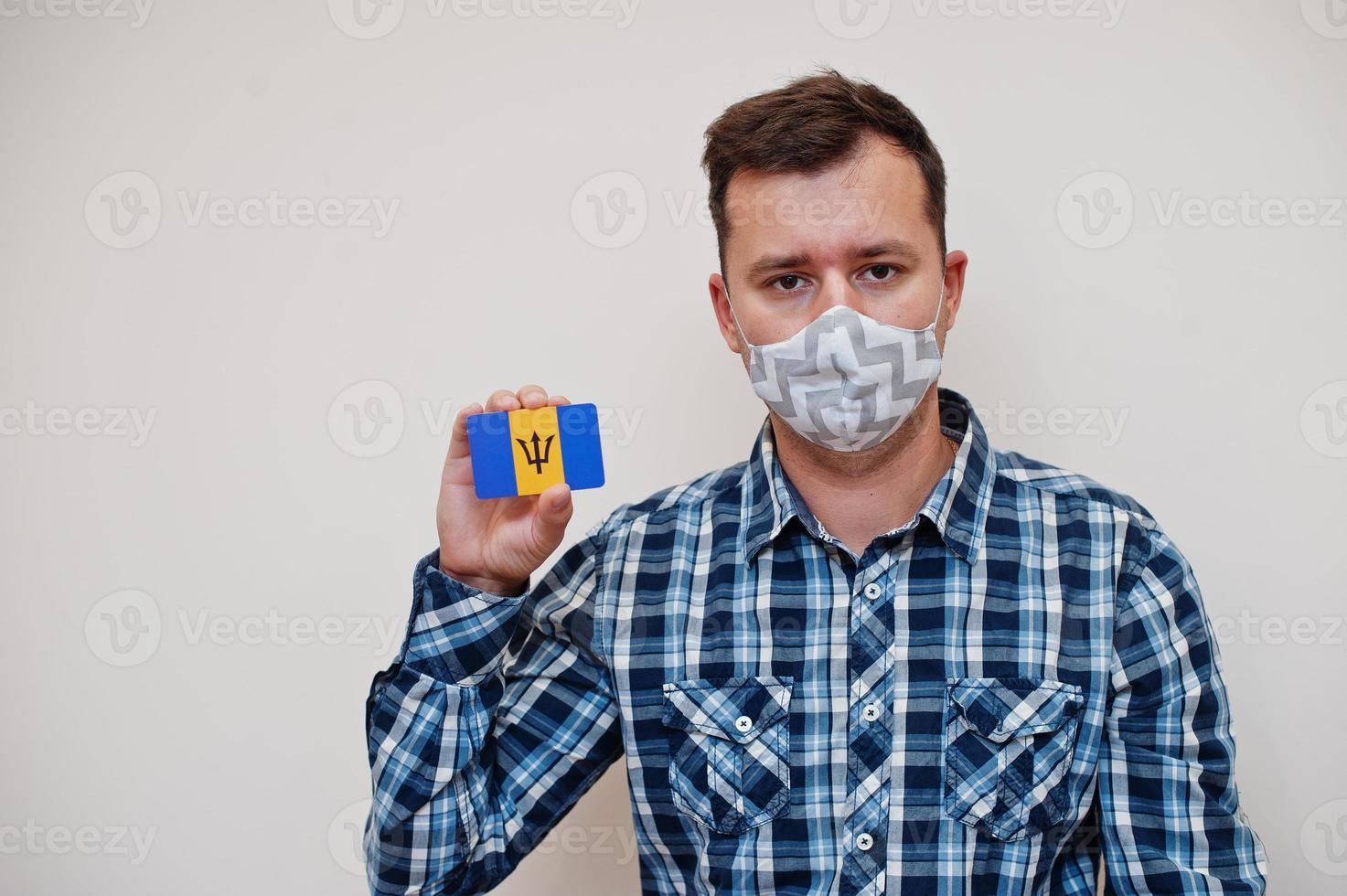 Man in checkered shirt show Barbados flag card in hand, wear protect mask isolated on white background. American countries Coronavirus concept. photo