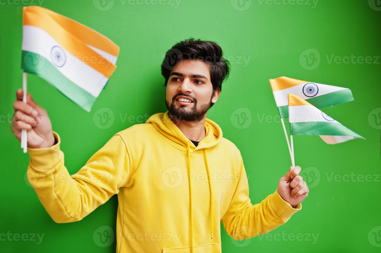 un joven indio hipster urbano con una sudadera amarilla de moda sostiene banderas de la india en las manos. Un chico fresco del sur de Asia usa una sudadera con capucha aislada en un fondo verde. foto