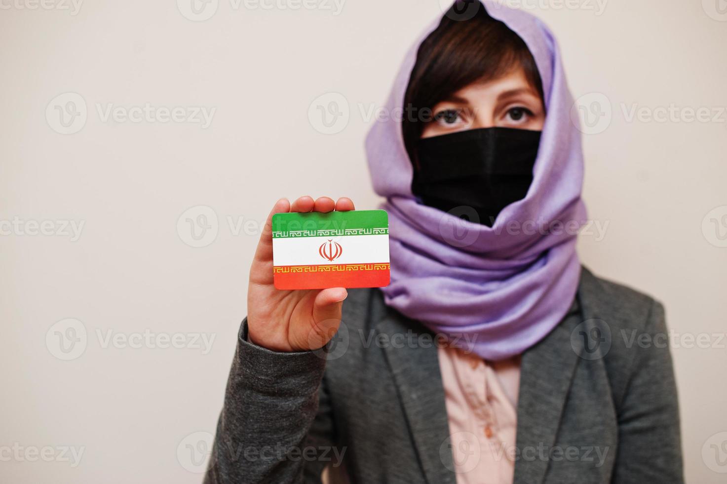 Portrait of young muslim woman wearing formal wear, protect face mask and hijab head scarf, hold Iran flag card against isolated background. Coronavirus country concept. photo