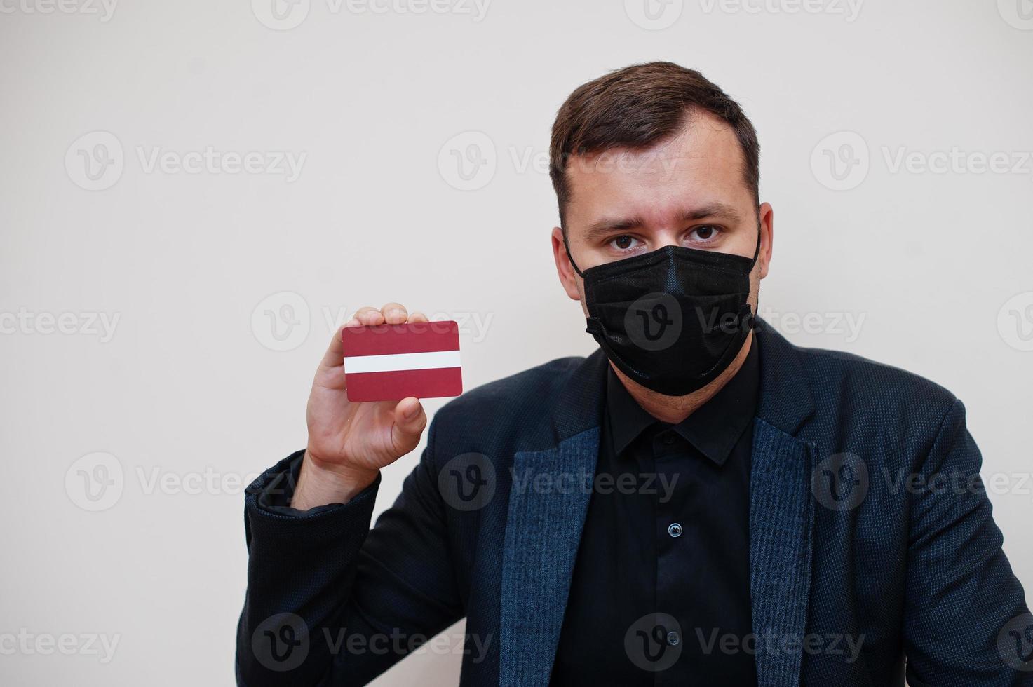 European man wear black formal and protect face mask, hold Latvia flag card isolated on white background. Europe coronavirus Covid country concept. photo