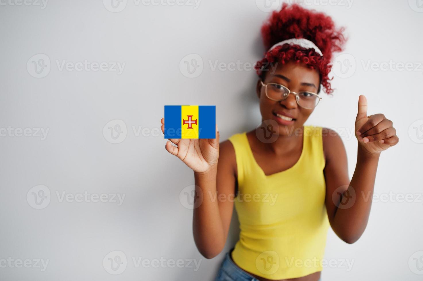 mujer africana con cabello afro, use camiseta amarilla y anteojos, sostenga la bandera de madeira aislada en fondo blanco, muestre el pulgar hacia arriba. foto