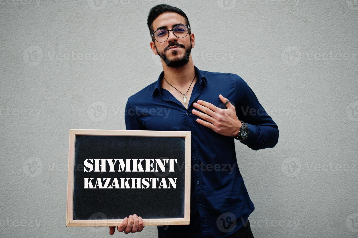 el hombre árabe usa camisa azul y anteojos sostiene el tablero con la inscripción shymkent kazakhstan. ciudades más grandes en el concepto del mundo islámico. foto