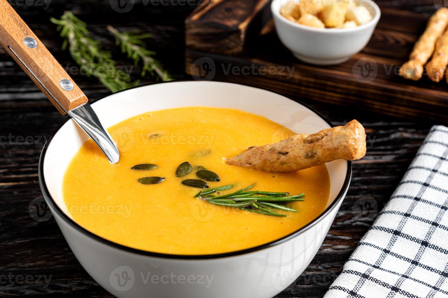 Pumpkin and carrot soup with cream, pumpkin seeds, croutons and grossini bread sticks on dar k wooden background. Rustic style, selective focus photo