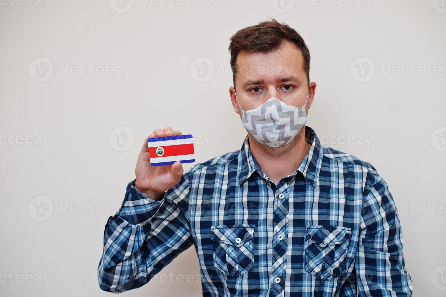 Man in checkered shirt show Costa Rica flag card in hand, wear protect mask isolated on white background. American countries Coronavirus concept. photo