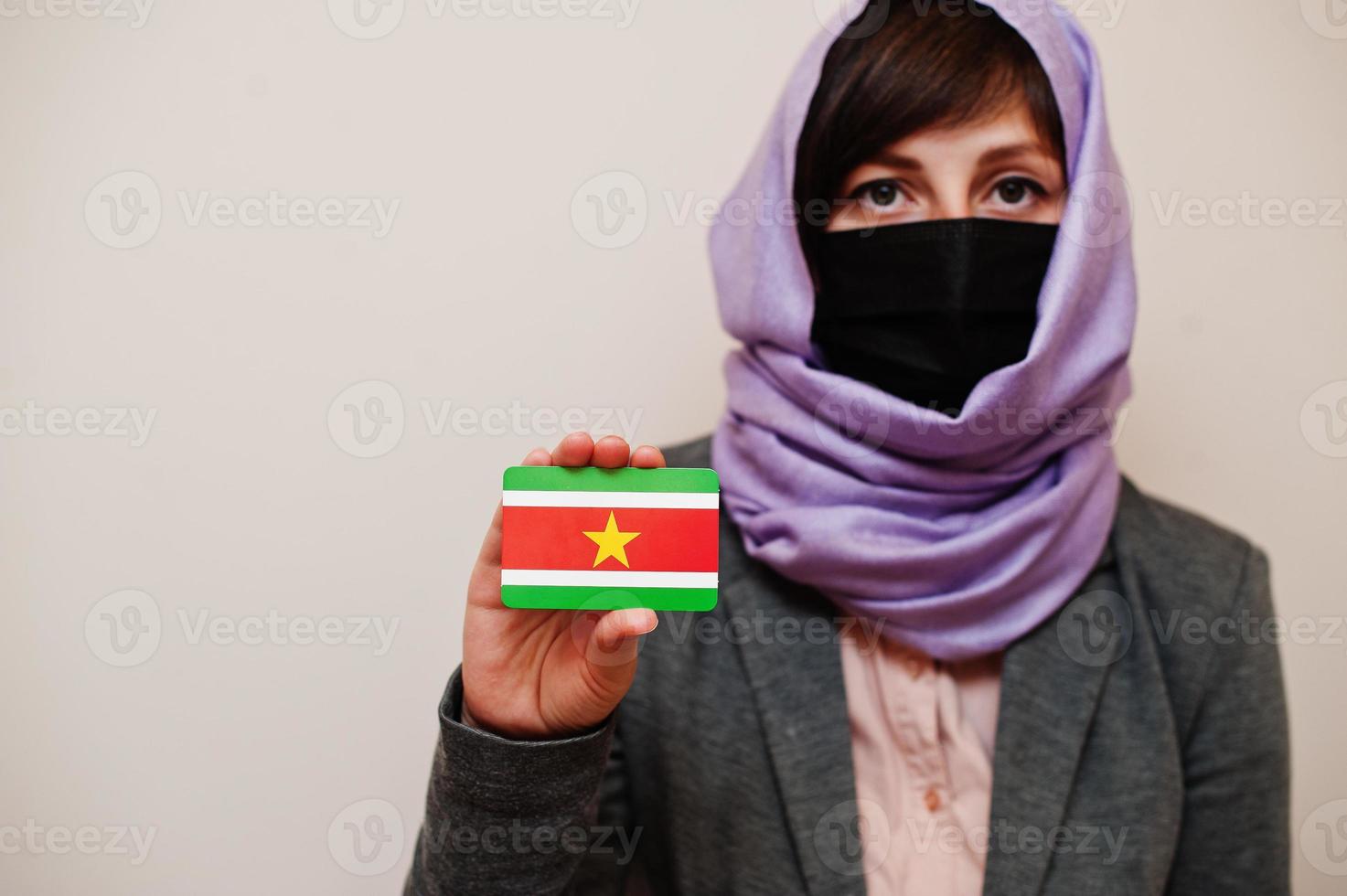 Portrait of young muslim woman wearing formal wear, protect face mask and hijab head scarf, hold Suriname flag card against isolated background. Coronavirus country concept. photo