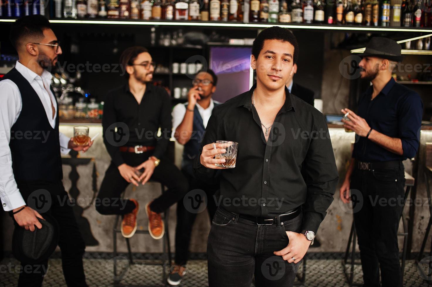 Stylish arab man against group of handsome retro well-dressed guys gangsters spend time at club, drinking on bar counter. Multiethnic male bachelor mafia party in restaurant. photo