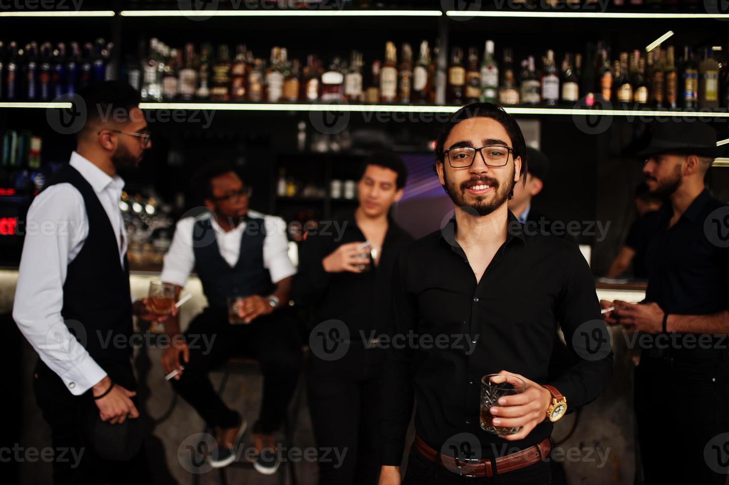 Stylish arab man against group of handsome retro well-dressed guys gangsters spend time at club, drinking on bar counter. Multiethnic male bachelor mafia party in restaurant. photo