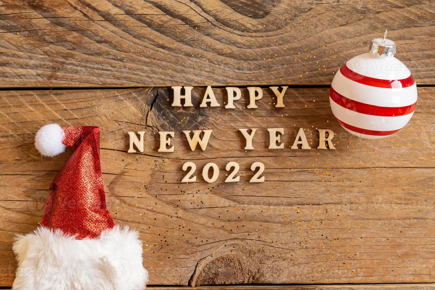 Happy new year 2022 wooden letters quote with red sparkling santa hat and striped bauble on wooden table. Festive greeting card for a new year celebration photo