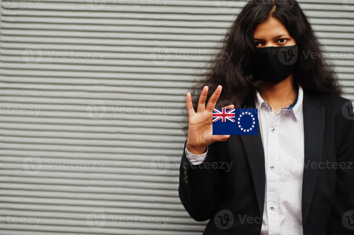 Asian woman at formal wear and black protect face mask hold Cook Islands flag at hand against gray background. Coronavirus at country concept. photo