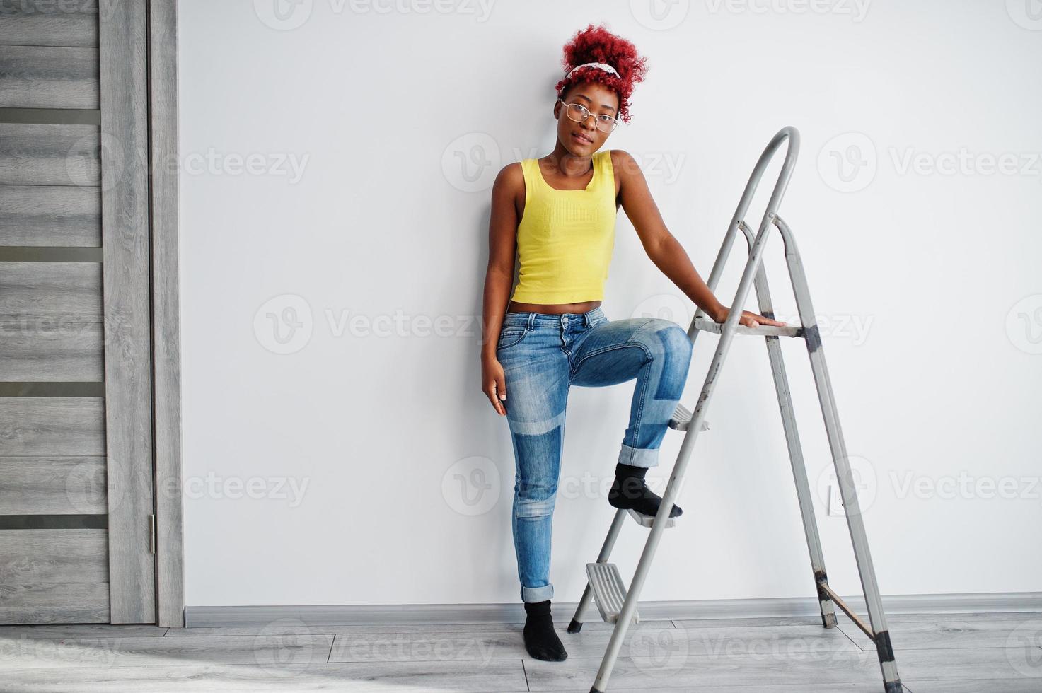 African american woman in yellow singlet and eyglasses against white wall stand om iron ladder. photo
