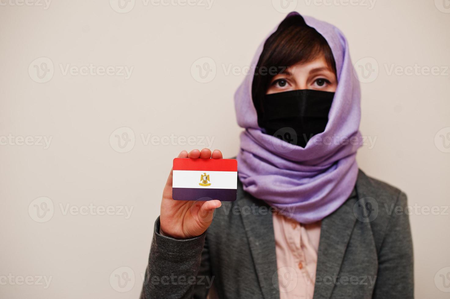 Portrait of young muslim woman wearing formal wear, protect face mask and hijab head scarf, hold Egypt flag card against isolated background. Coronavirus country concept. photo