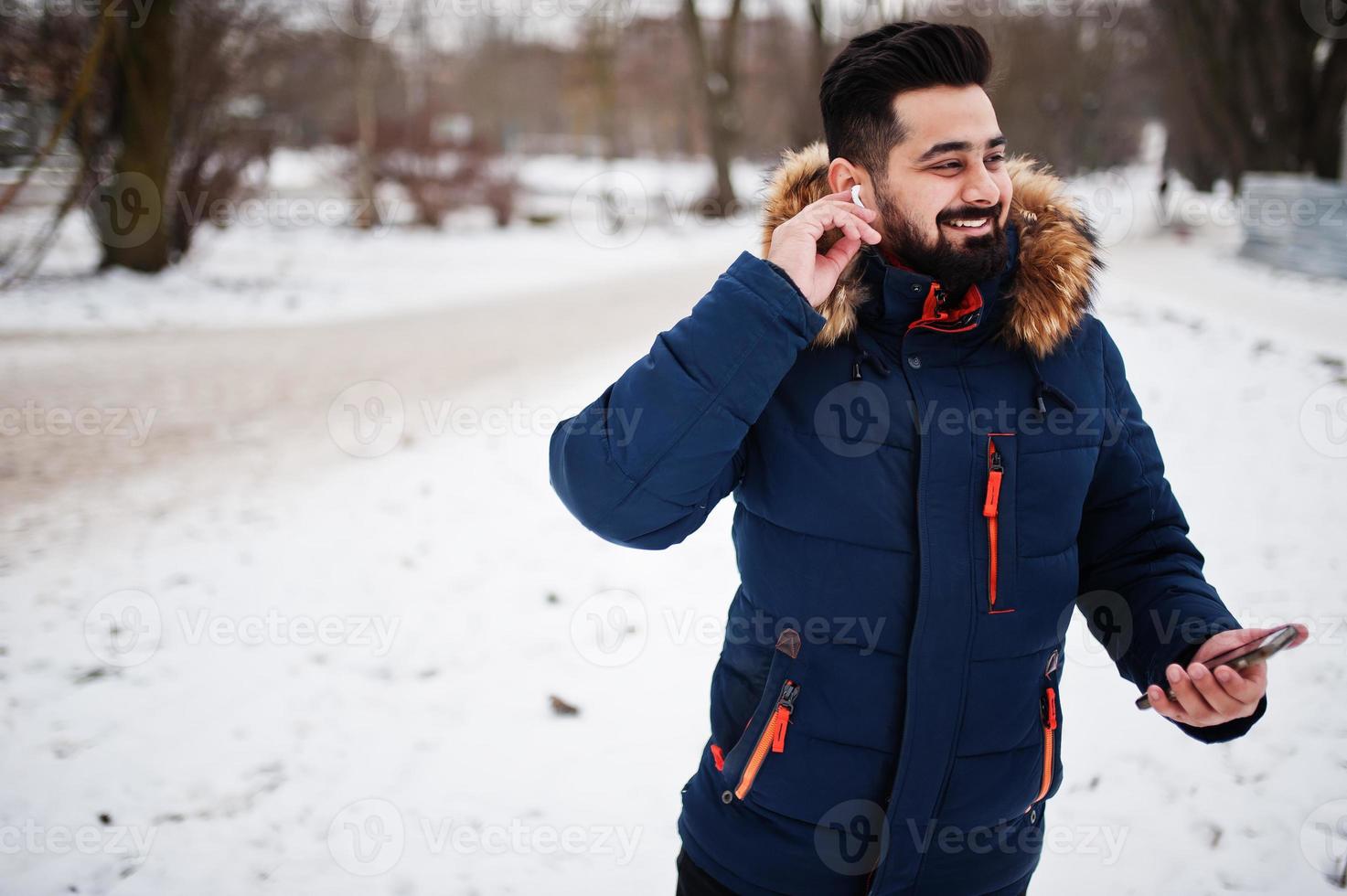 Beard indian man wear jacket in cold winter day. He speak mobile phone by wireless headphones. photo