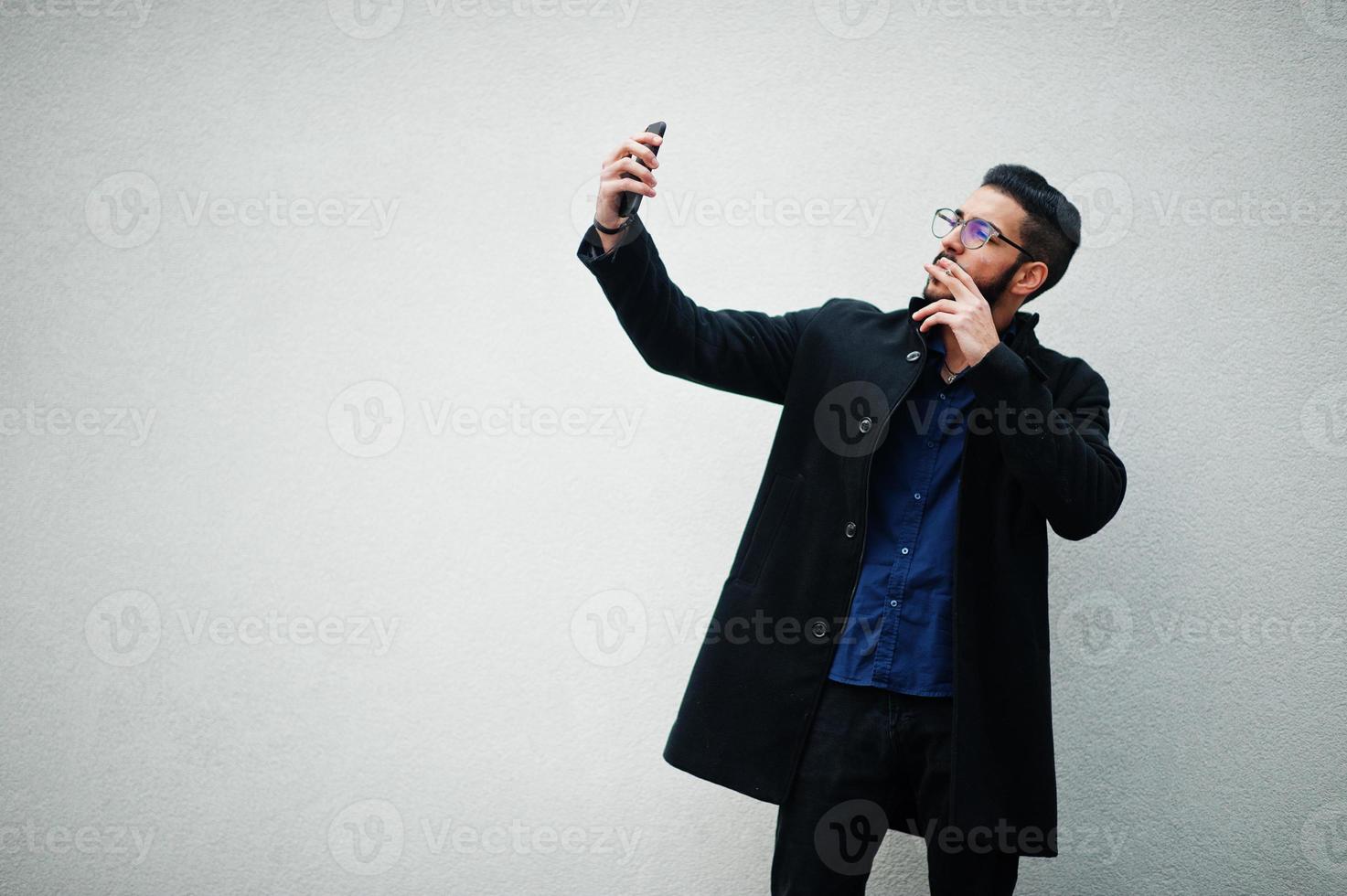 Middle eastern entrepreneur wear black coat and blue shirt, eyeglasses standing against white wall, smoking cigarette and making selfie by phone. photo