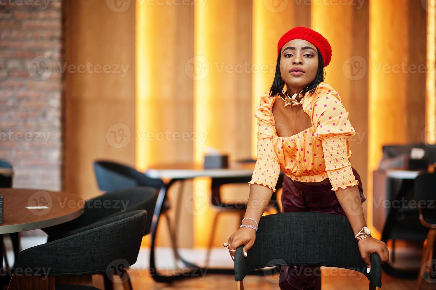 Elegant afro american woman in red french beret, big gold neck chain polka dot blouse and leather pants pose indoor. photo