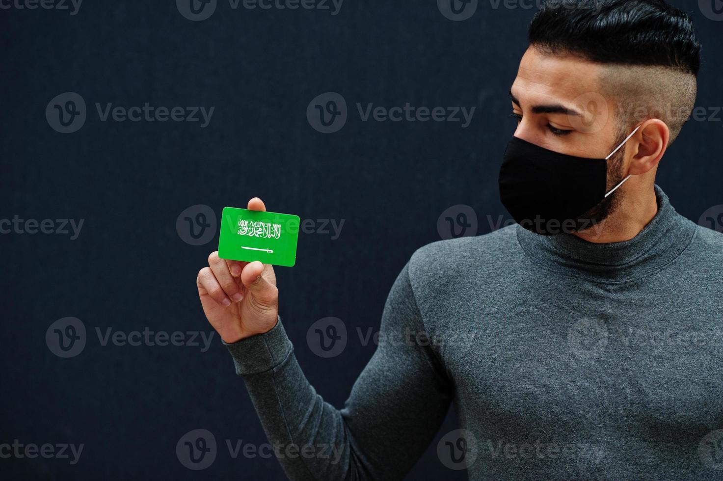 hombre de oriente medio con cuello de tortuga gris y máscara protectora de cara negra muestra antecedentes aislados de la bandera de arabia saudita. foto