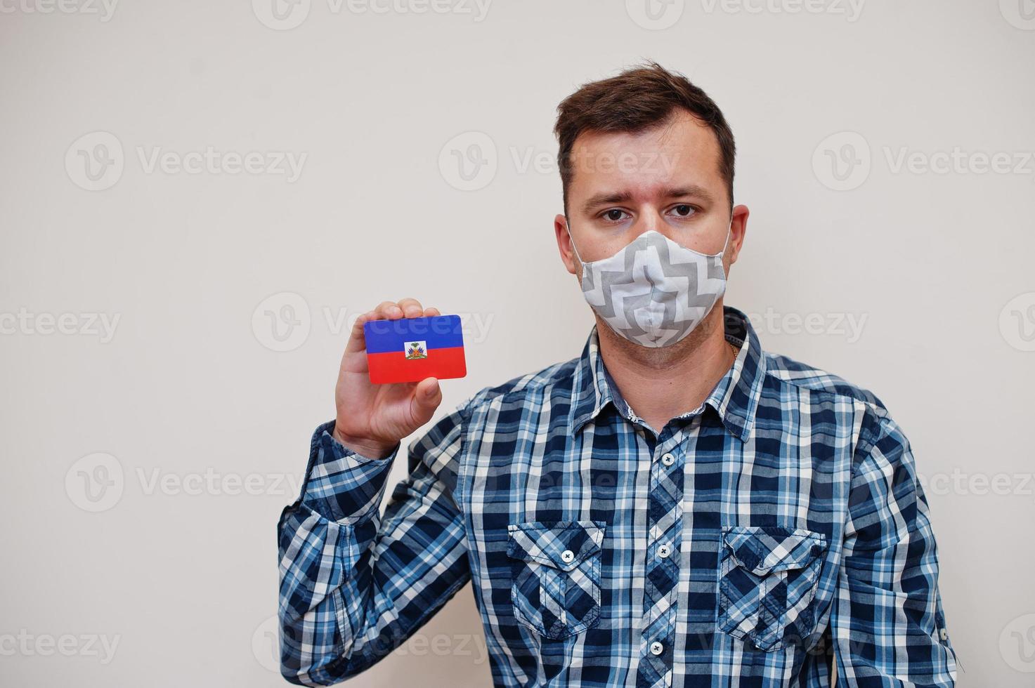 Man in checkered shirt show Haiti flag card in hand, wear protect mask isolated on white background. American countries Coronavirus concept. photo