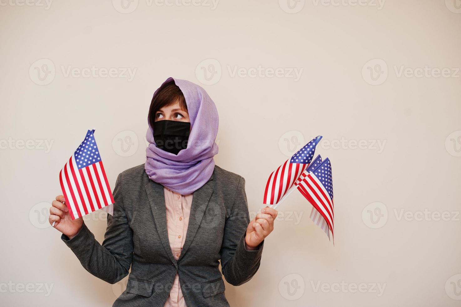 Portrait of young muslim woman wearing formal wear, protect face mask and hijab head scarf, hold USA flags in hands against isolated background. Coronavirus country concept. photo