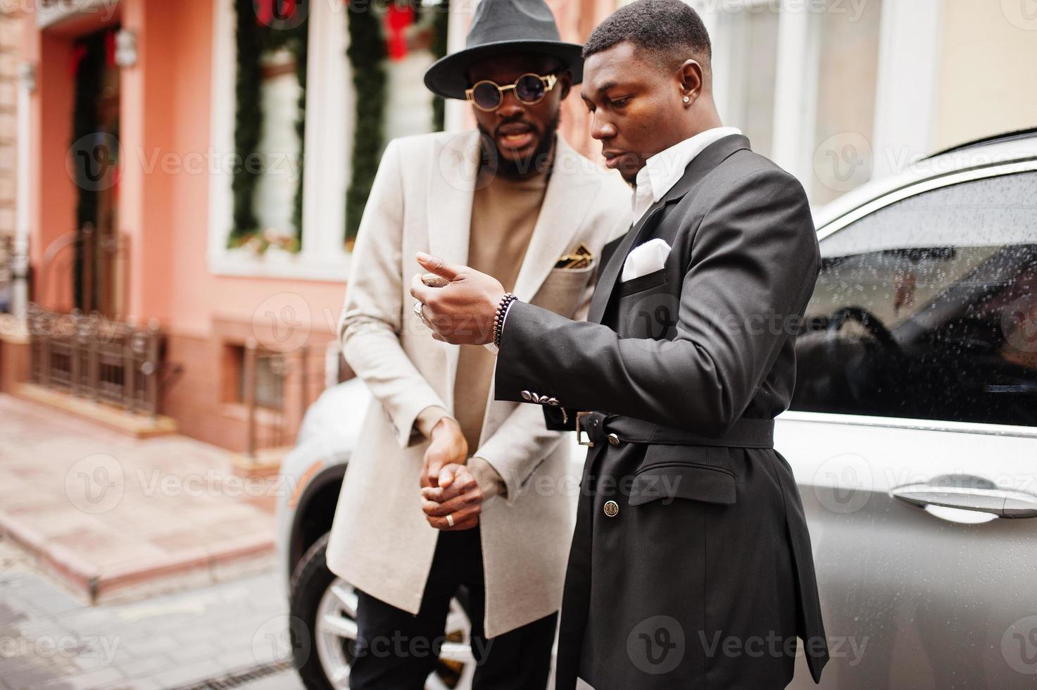 dos hombres negros de moda se paran cerca del auto de negocios y miran el teléfono celular. retrato de moda de modelos masculinos afroamericanos. llevar traje, abrigo y sombrero. foto