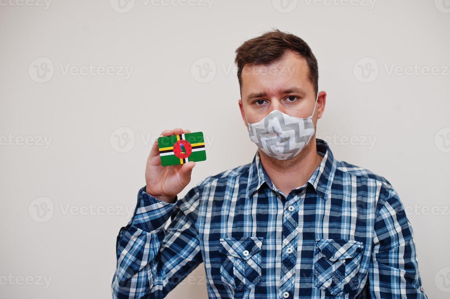Man in checkered shirt show Dominica flag card in hand, wear protect mask isolated on white background. American countries Coronavirus concept. photo