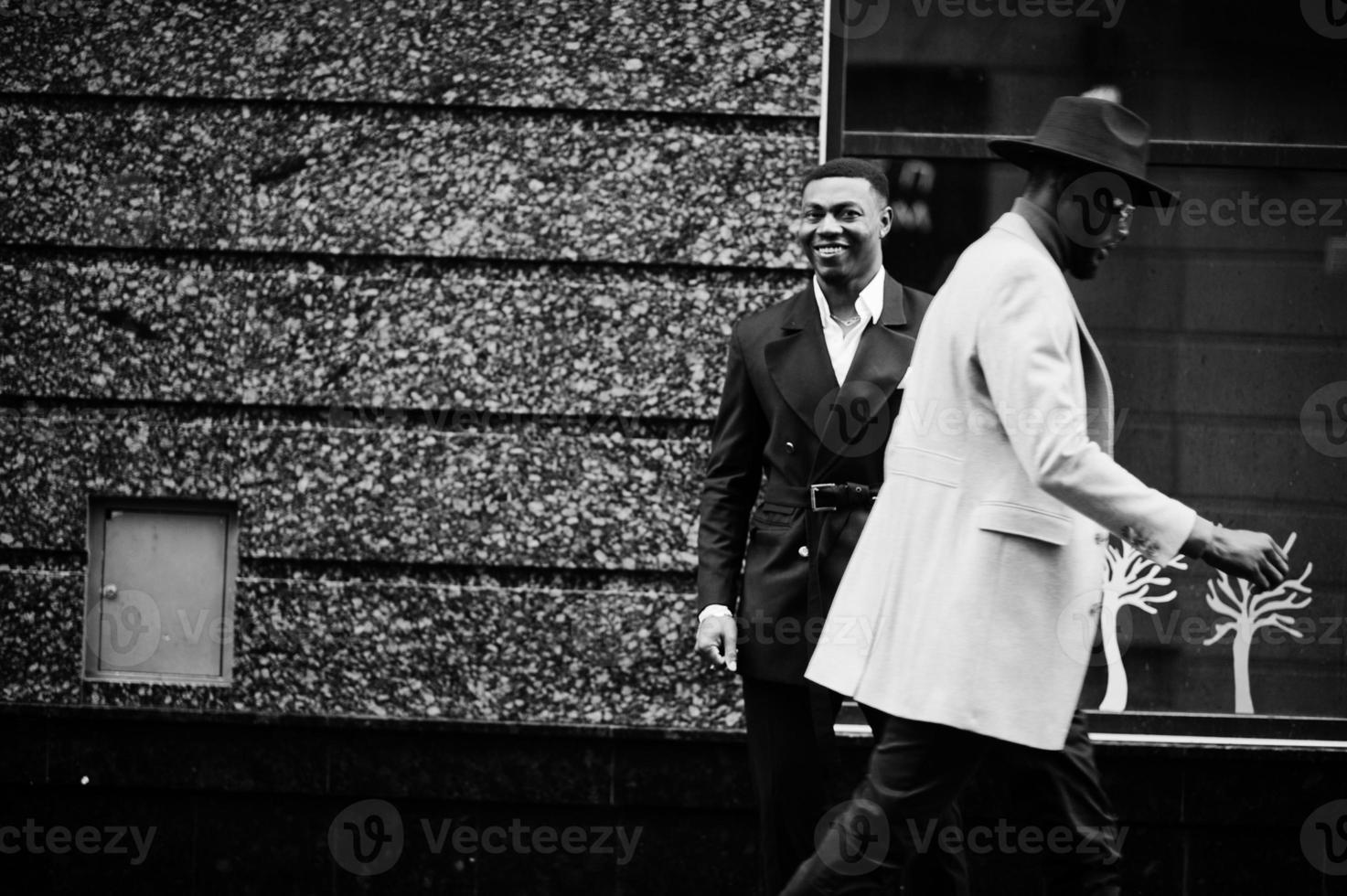 Two fashion black men walking on street. Fashionable portrait of african american male models. Wear suit, coat and hat. photo