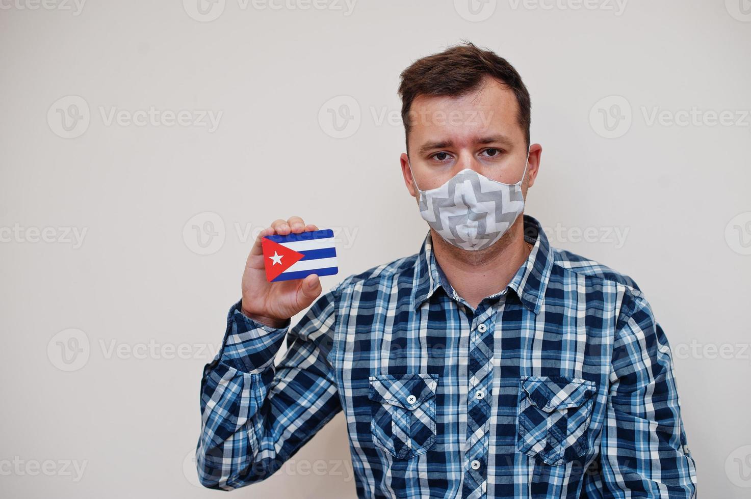 Man in checkered shirt show Cuba flag card in hand, wear protect mask isolated on white background. American countries Coronavirus concept. photo