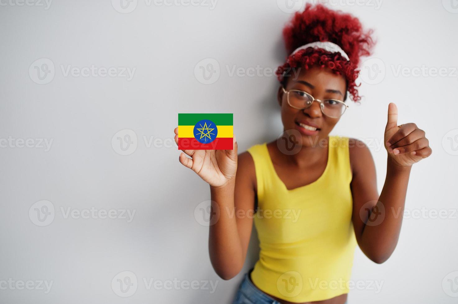 African woman with afro hair, wear yellow singlet and eyeglasses, hold Ethiopia flag isolated on white background, show thumb up. photo