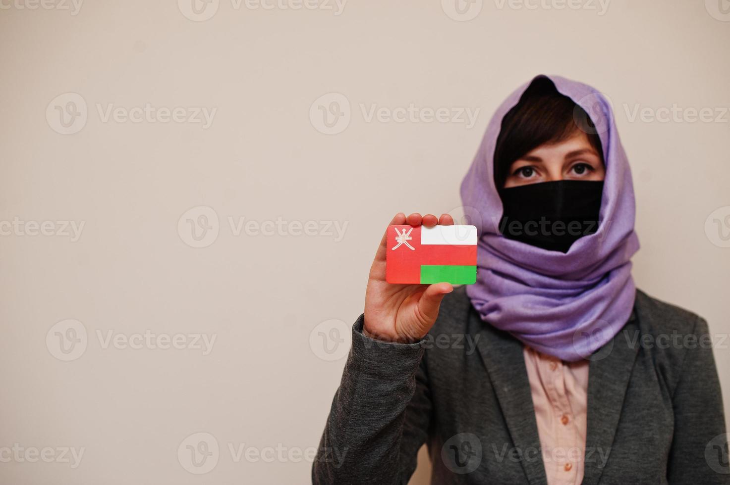 Portrait of young muslim woman wearing formal wear, protect face mask and hijab head scarf, hold Oman flag card against isolated background. Coronavirus country concept. photo