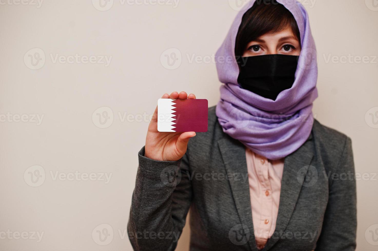 Portrait of young muslim woman wearing formal wear, protect face mask and hijab head scarf, hold Qatar flag card against isolated background. Coronavirus country concept. photo