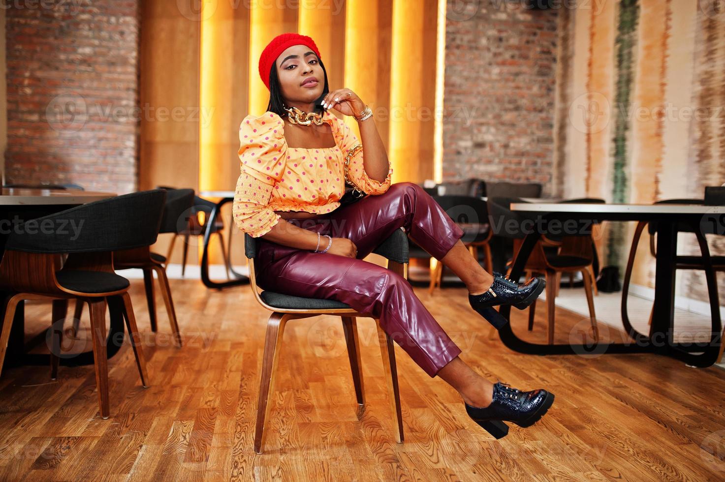 elegante mujer afroamericana con boina francesa roja, blusa de lunares con cadena de cuello dorado grande y pantalones de cuero posan en el interior. foto