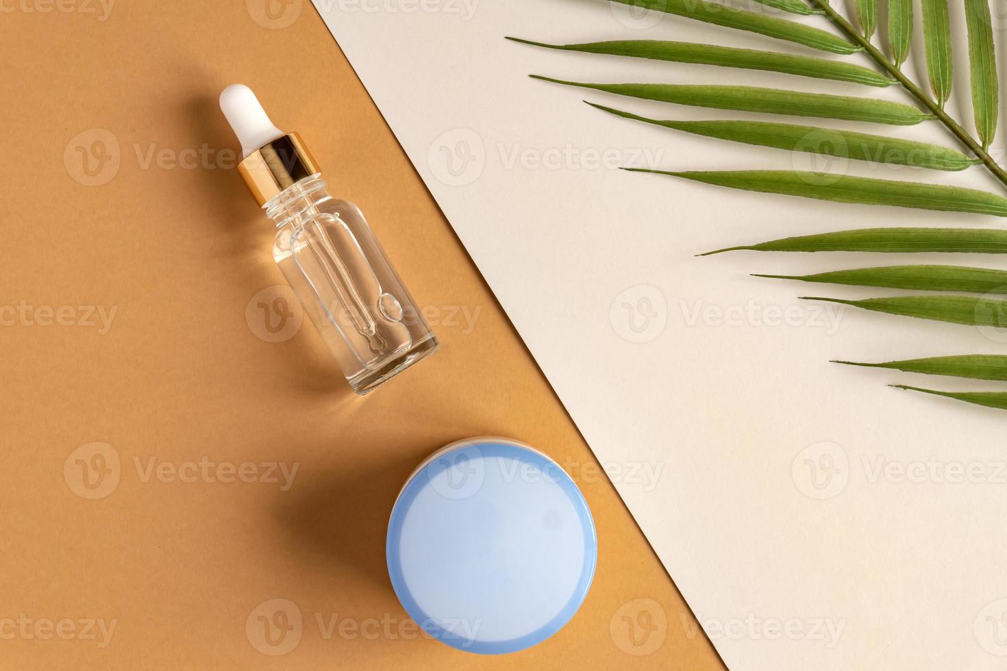 anti-aging collagen facial serum in transparent glass bottle with gold pipette and face cream on double beige and gray background decorated with stropical branch. Organic Cosmetic Concept photo
